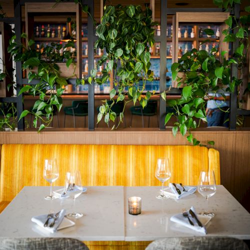 A restaurant table set for four with wine glasses, napkins, and a candle, surrounded by green plants and modern decor, ambient lighting.