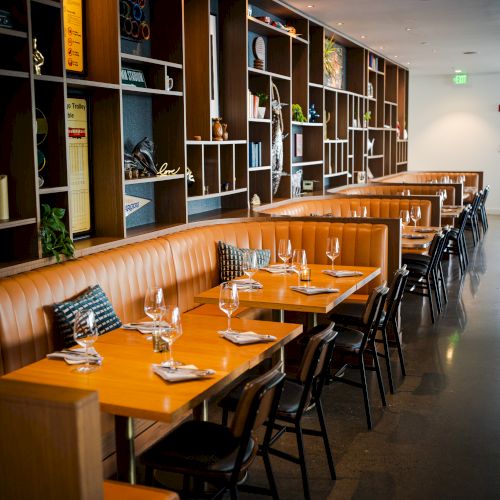 A modern restaurant with wooden tables, leather booths, and neatly set dining arrangements, featuring shelves with decorative items on the wall.