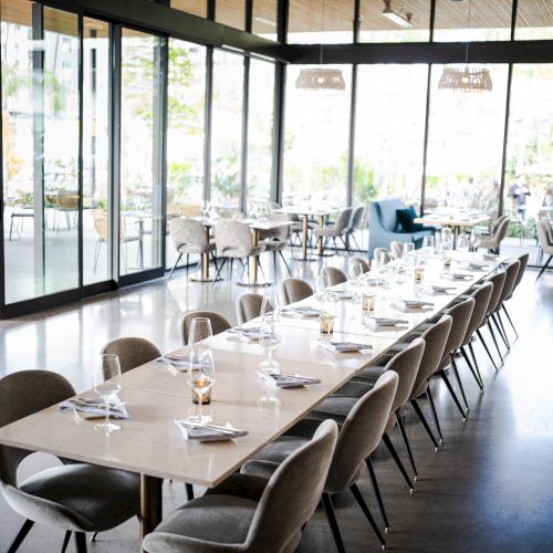 The image shows a modern, well-lit dining area with large glass windows, featuring a long table set with glassware and surrounded by multiple chairs.