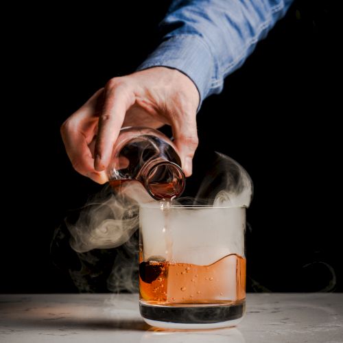 A hand pours a dark liquid into a glass with a smoky effect, possibly dry ice or a similar element adding visual intrigue to the drink.