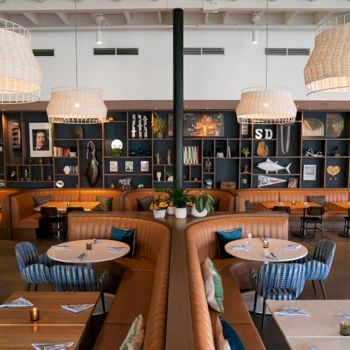 Stylish restaurant interior with cozy booths, round tables, decorative shelves, and large hanging lamps. Walls feature various artworks.