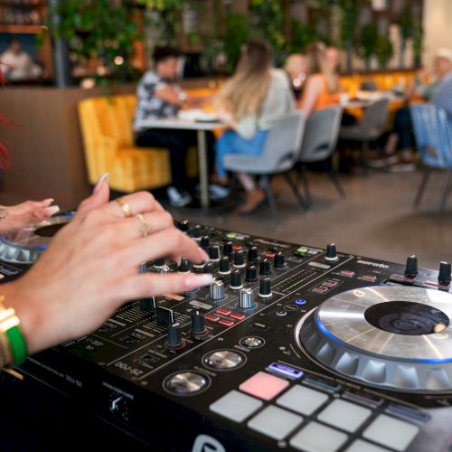 A DJ is seen mixing music using a console in a lively indoor setting, with people socializing and dining in the background.