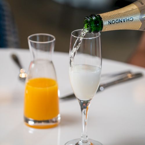 A champagne bottle is pouring champagne into a flute next to a carafe of orange juice on a white table with utensils in the background.