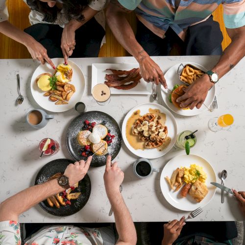 Four people enjoying brunch at a table with plates of pancakes, toast, bacon, eggs, coffee, and juice drinks, viewed from above.