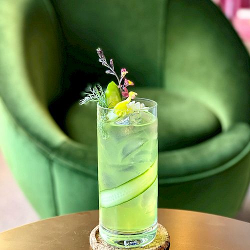 A tall glass of a green drink with ice, garnished with herbs and lime, sits on a coaster atop a wooden table in front of a green chair.