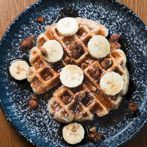 A plate with a waffle topped with banana slices, powdered sugar, and syrup is on a wooden table.