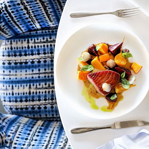 A restaurant table with a dish of seared tuna and colorful vegetables, alongside a wine glass, cutlery, and a blue patterned chair.