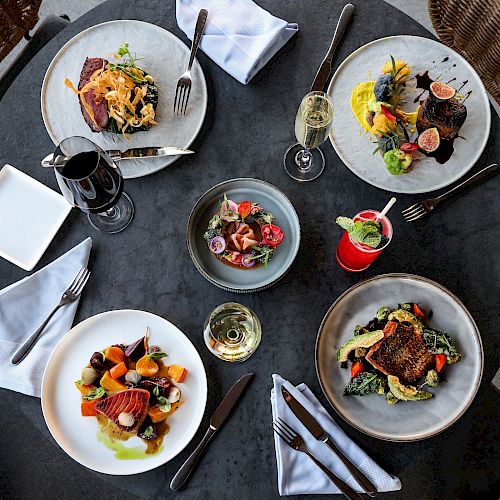 An overhead view of a table with four gourmet dishes, drinks, cutlery, and napkins set neatly on a grey table, served in an upscale setting.