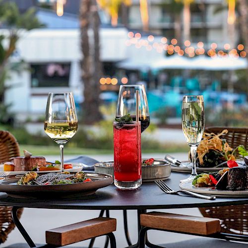 A table set for two with diverse dishes, two glasses of white wine, and a red cocktail, in an outdoor setting with decorative lights.