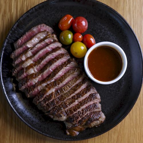 The image shows a sliced steak on a black plate, accompanied by cherry tomatoes and a small bowl of sauce on a wooden surface.