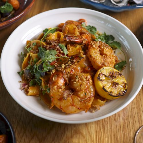 A bowl of shrimp pasta garnished with lemon and herbs is set on a table among other dishes and glasses of white wine.