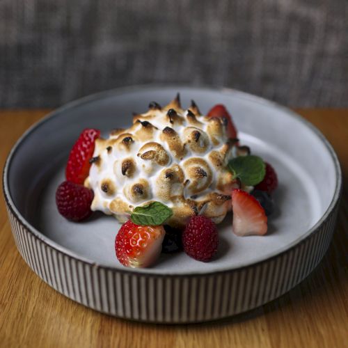 A gourmet dessert with toasted meringue, surrounded by strawberries and raspberries, garnished with fresh mint leaves, served in a white bowl.