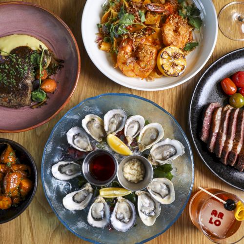 This image shows a variety of dishes including oysters, steak, shrimps, and sides, along with a drink and condiments, all arranged on a wooden table.