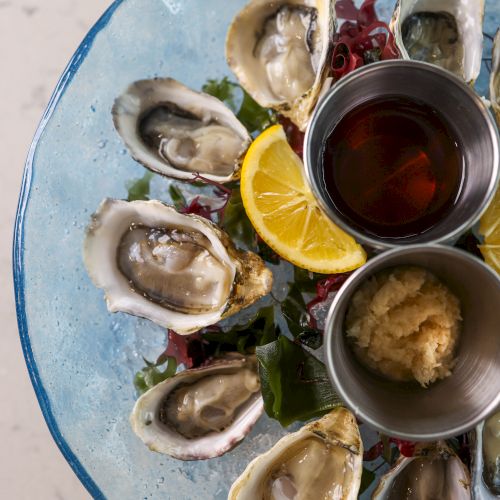 The image shows a plate of fresh oysters with lemon slices, sauce, and a condiment in small containers garnished with what appears to be seaweed.