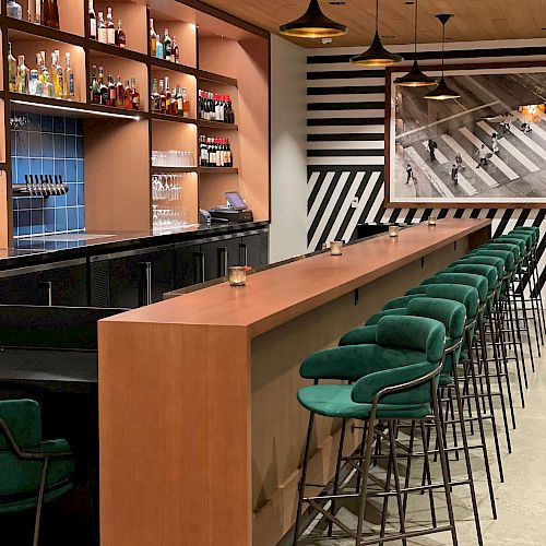 A modern bar with green high stools lined up along a wooden counter, shelves of bottles, pendant lights, and striped wall art in the background.