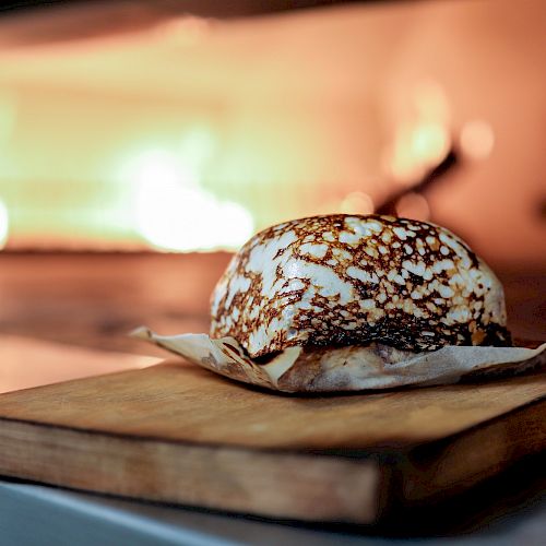 This image shows a baked item with a browned top on parchment paper, placed on a wooden board, in front of an open flame oven.