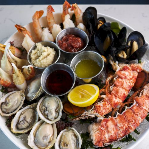 The image shows a seafood platter with shrimp, oysters, mussels, lobster tail, and various dipping sauces, garnished with lemon slices.