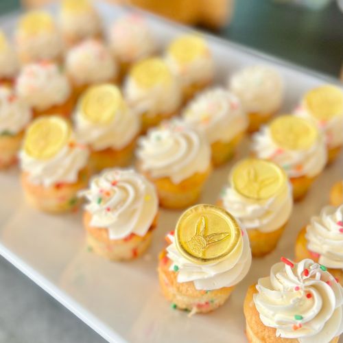 Mini cupcakes with white frosting, colorful sprinkles, and yellow decorations on top are arranged on a tray.