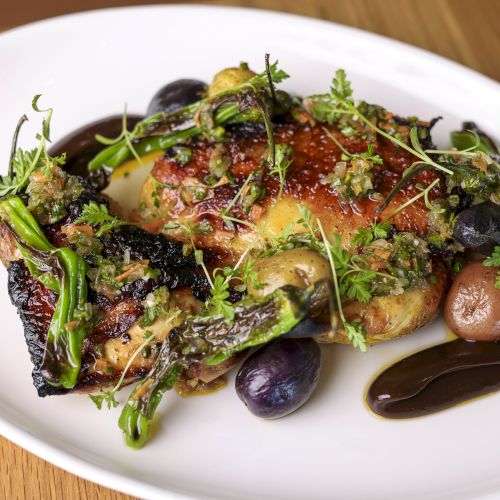 The image shows a plate with a gourmet dish featuring roasted chicken, garnished with greens and purple grapes, served with a dark sauce.