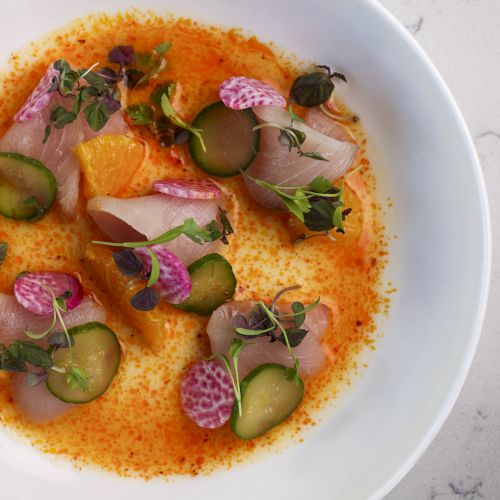 A white bowl with a gourmet dish featuring sliced fish, microgreens, cucumbers, radish, and an orange sauce on a light gray speckled surface.