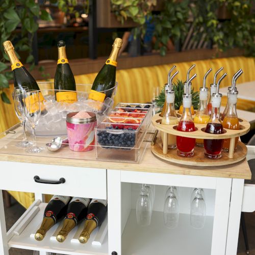 Champagne bottles in ice, glasses, bowls of fruit, flavor bottles on a cart in a dining area with green plants and yellow seating.