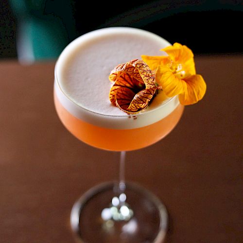 A cocktail in a glass garnished with a dried citrus slice and a yellow flower on a brown surface.