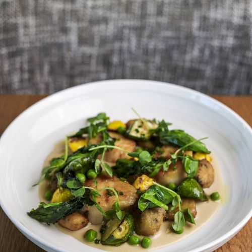 A white bowl filled with a dish comprising gnocchi, green vegetables, and garnished with fresh herbs sits on a wooden table, with a chair in the background.