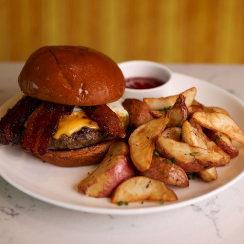 A hamburger with melted cheese and crispy bacon, served with potato wedges and a small dish of ketchup.