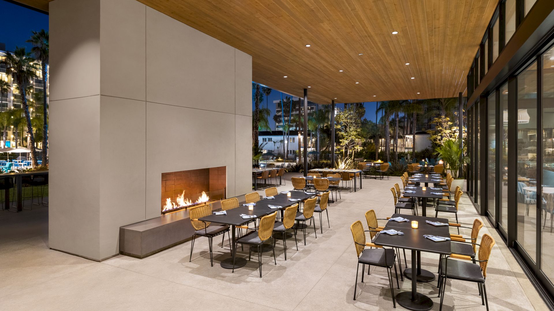 An outdoor dining area with modern decor features a fireplace, multiple tables, and chairs under a wooden ceiling at nighttime.