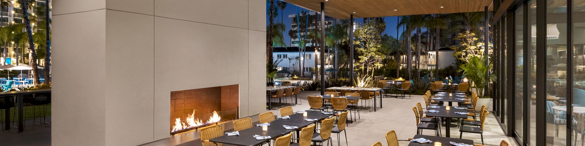 An outdoor dining area with modern decor features a fireplace, multiple tables, and chairs under a wooden ceiling at nighttime.