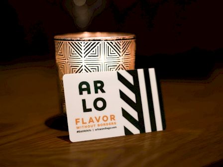 A lit candle holder with geometric patterns and a card labeled "ARLO FLAVOR WITHOUT BORDERS" placed in front of it on a wooden surface.