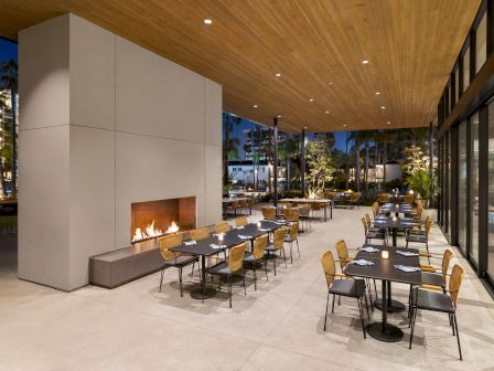 This image shows an outdoor dining area with a large fireplace, multiple tables and chairs, under a wooden roof, with a view of a nighttime scene.