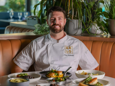 A person in a chef's uniform sits at a table with various dishes and plants in the background.