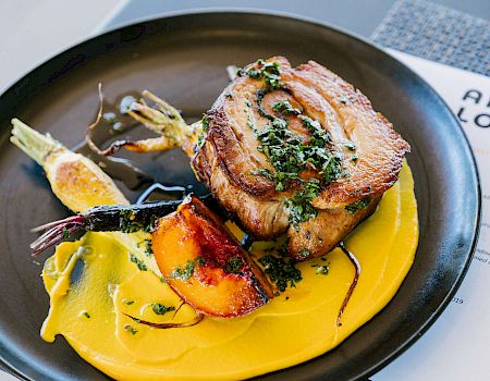 A plated dish featuring roasted meat, colorful carrots, and squash puree is garnished with herbs and paired with a restaurant menu in the background.