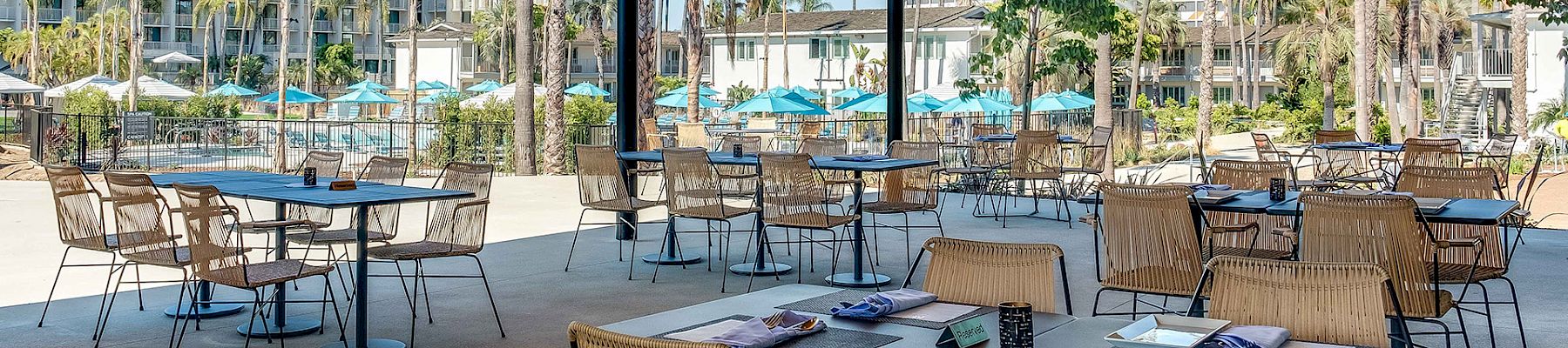 Outdoor dining area with several tables and chairs, set against a backdrop of palm trees and a pool area with blue umbrellas.