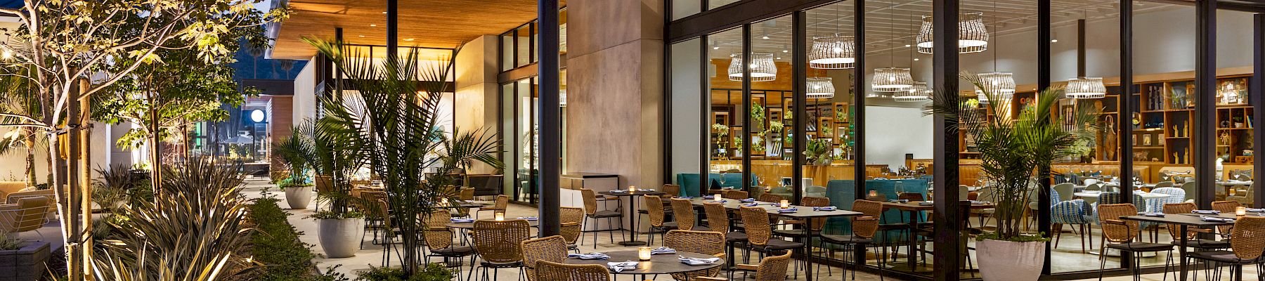 An outdoor restaurant patio features numerous tables and chairs, surrounded by potted plants and modern architecture under a wooden overhang, at dusk.