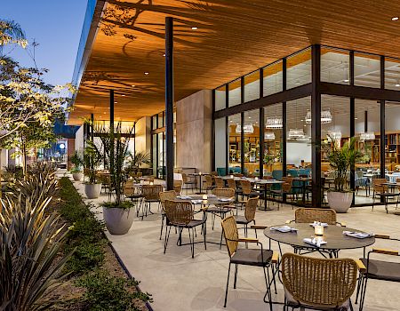 An outdoor restaurant patio features numerous tables and chairs, surrounded by potted plants and modern architecture under a wooden overhang, at dusk.