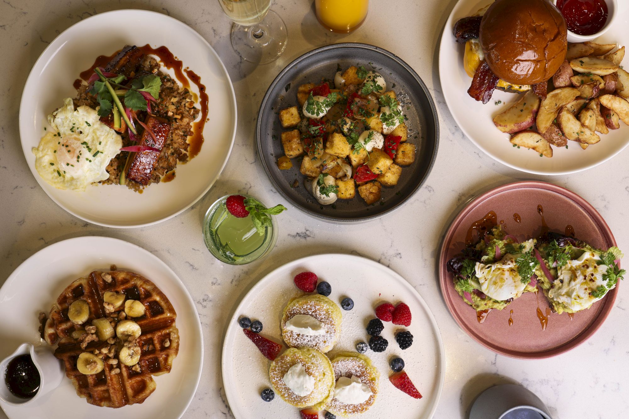 The image shows a variety of brunch dishes, including waffles with banana, eggs benedict, stuffed donuts, mixed berries, roasted potatoes, and a sandwich.