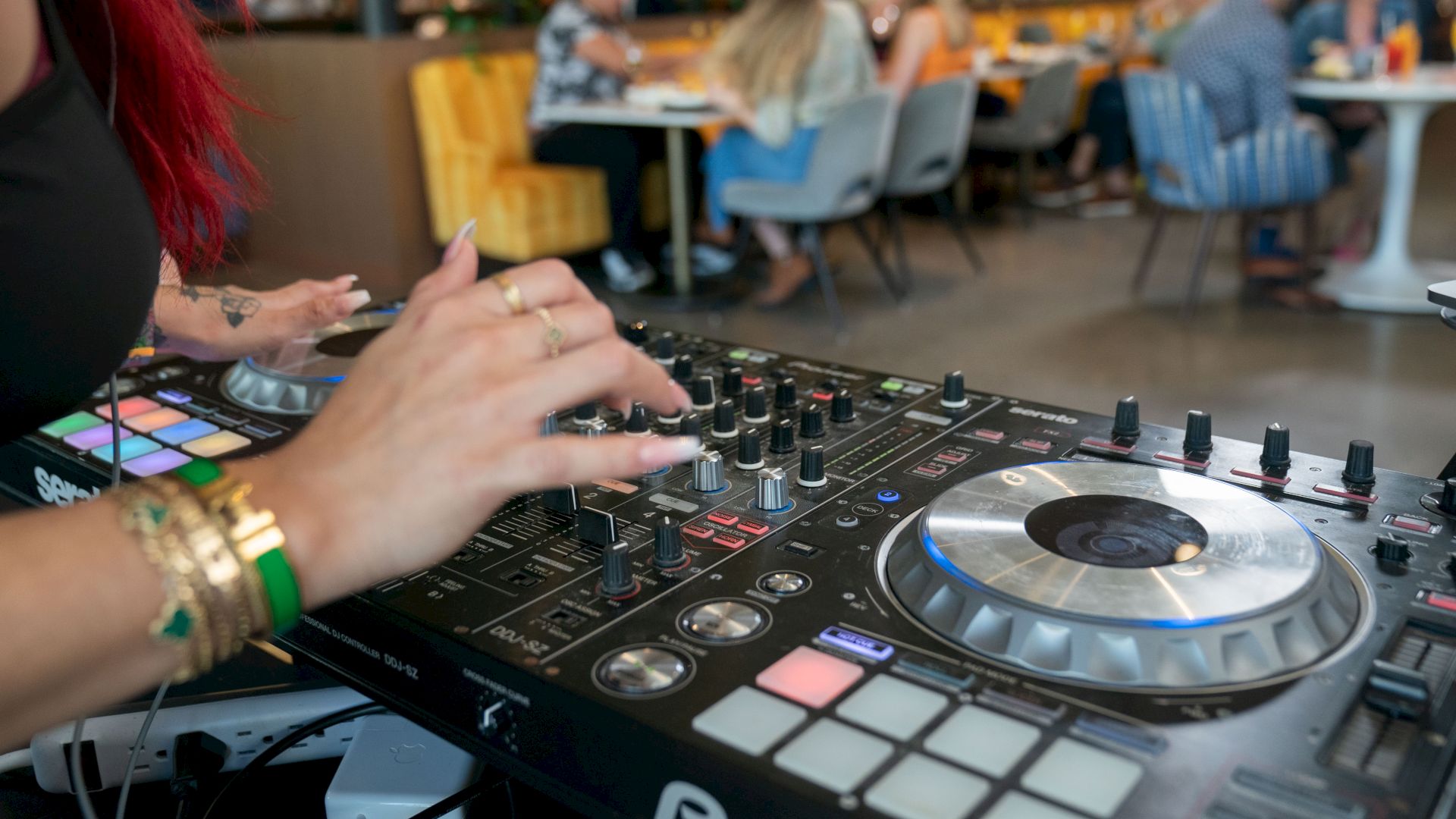 A DJ is mixing music on a console with a lively restaurant scene in the background, showcasing a social and vibrant atmosphere.