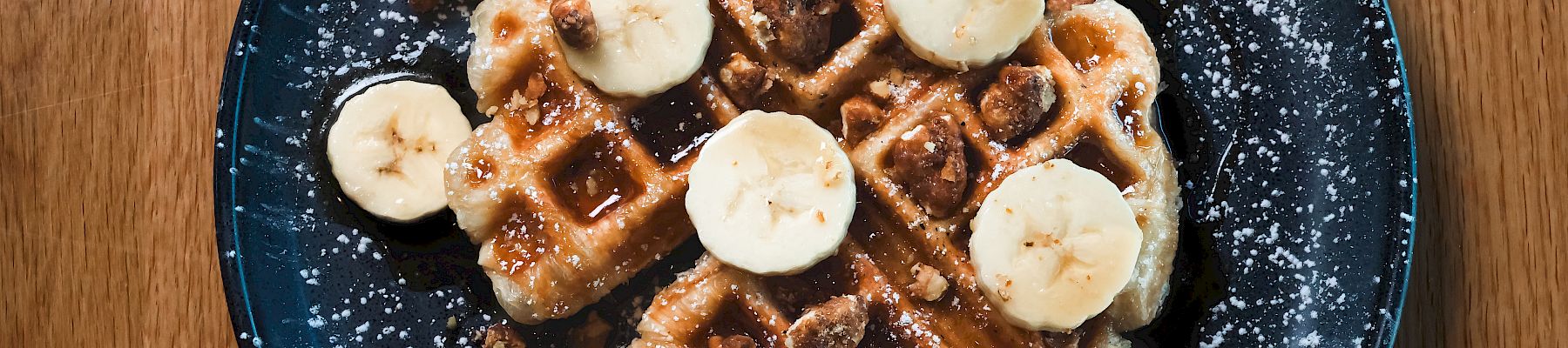 A plate with waffles topped with banana slices, powdered sugar, and syrup on a dark blue plate on a wooden table.