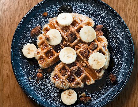 A plate with waffles topped with banana slices, powdered sugar, and syrup on a dark blue plate on a wooden table.