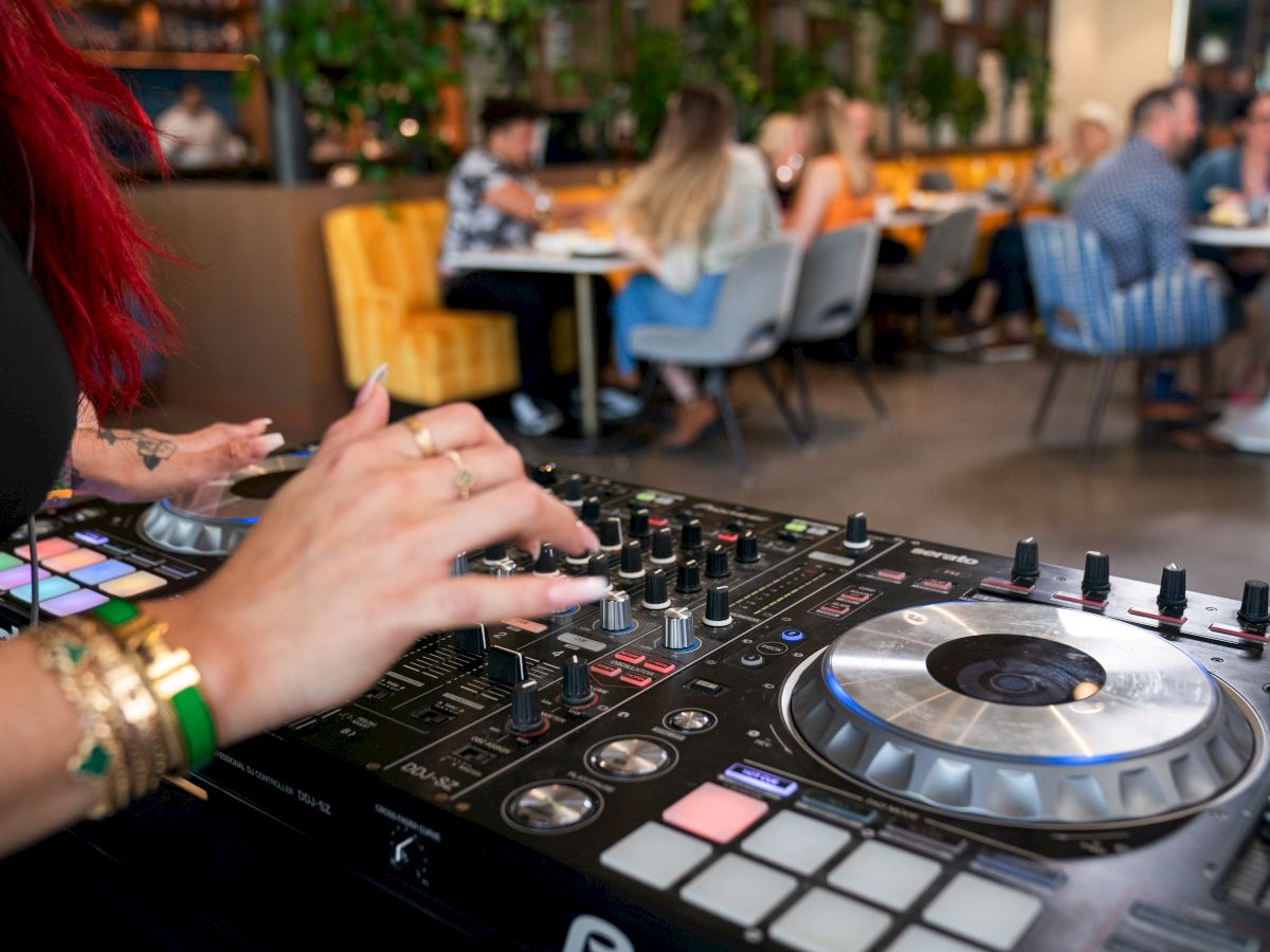 A DJ is mixing music on a console in a lively restaurant, with guests seated at tables in the background enjoying their time.