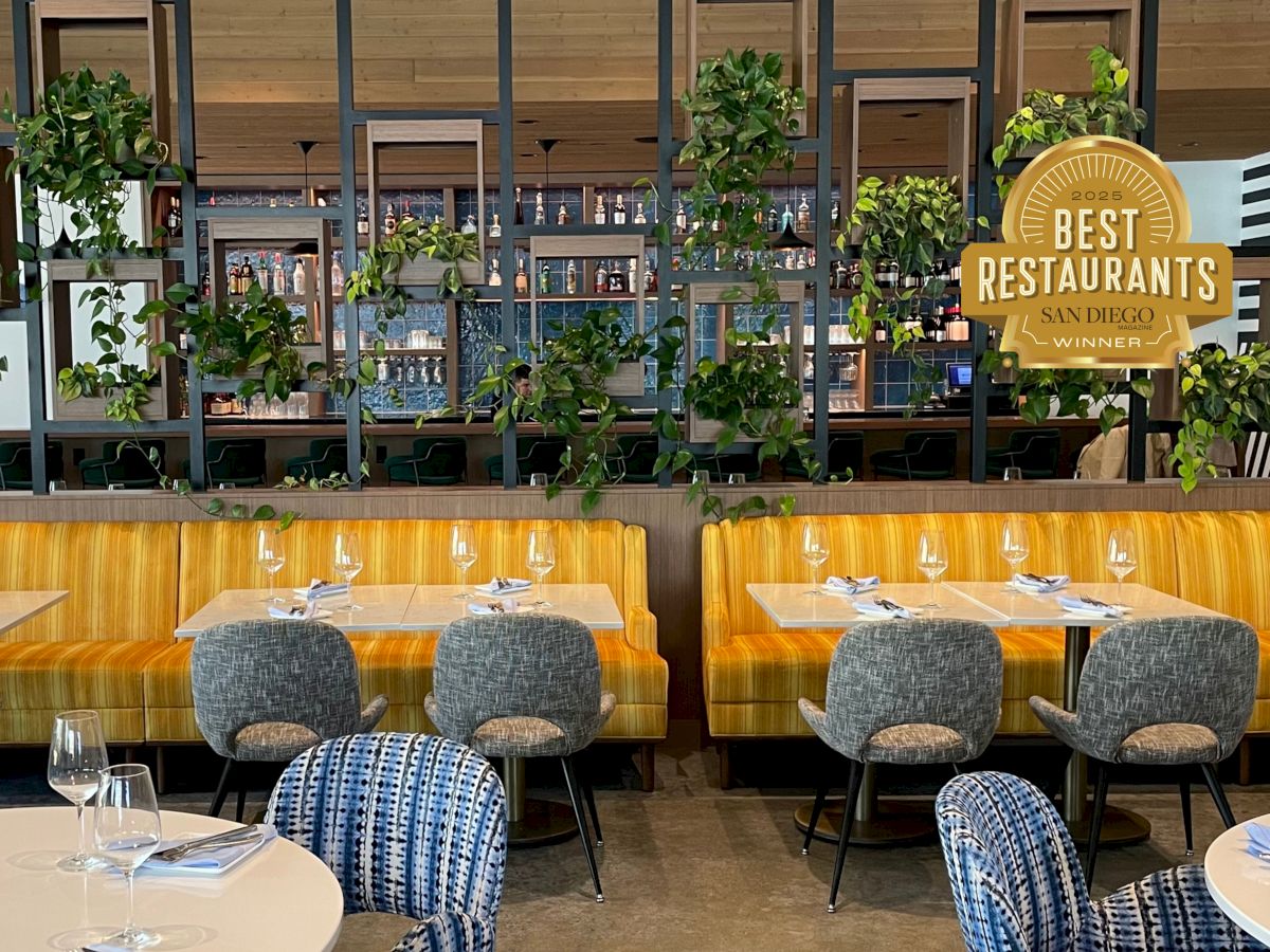 A stylish restaurant interior with yellow booths, gray chairs, a "Best Restaurants" award sign, and plants adorning a black grid.