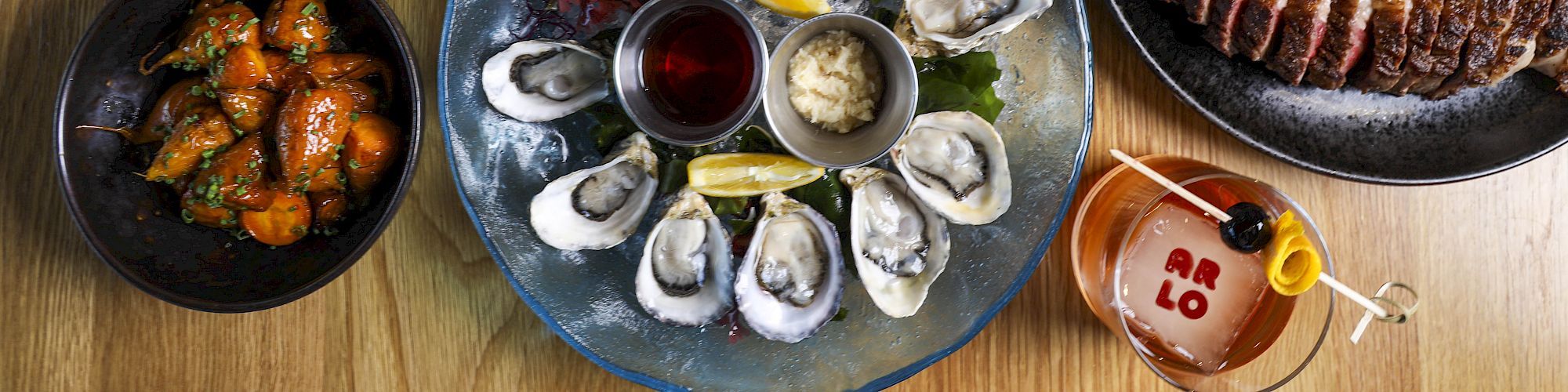 The image shows a table with various dishes, including oysters, sliced steak, grilled shrimp, chicken wings, and a cocktail.