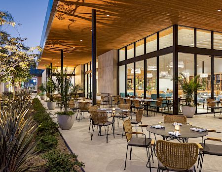 An outdoor dining area with several tables and chairs is adjacent to a modern glass-walled building, with lush plants and evening lighting enhancing the ambiance.