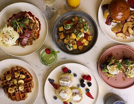 The image shows a table with plates of various foods, including waffles, eggs benedict, breakfast potatoes, a burger, and fresh fruits.