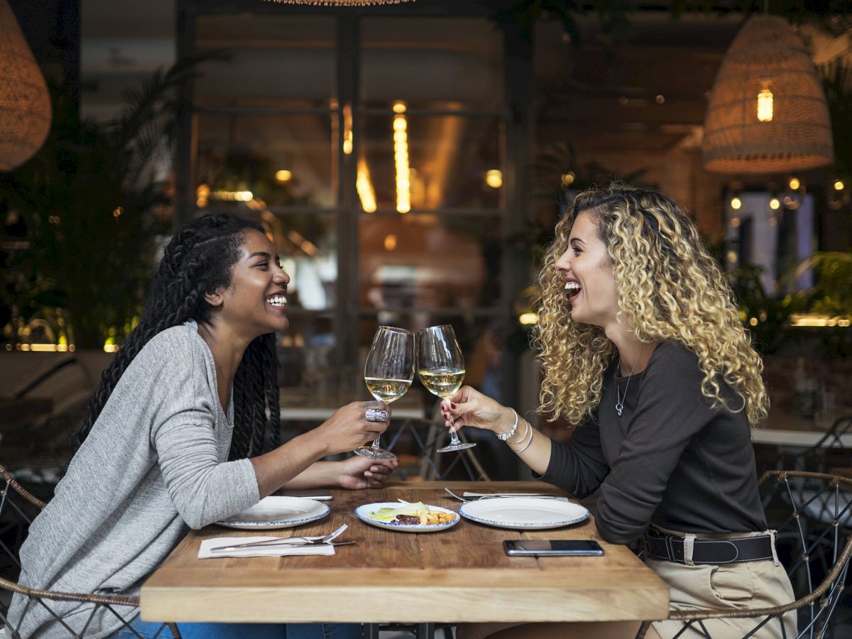 Two people are sitting at a restaurant table, clinking glasses of white wine and smiling, with plates and food in front of them.