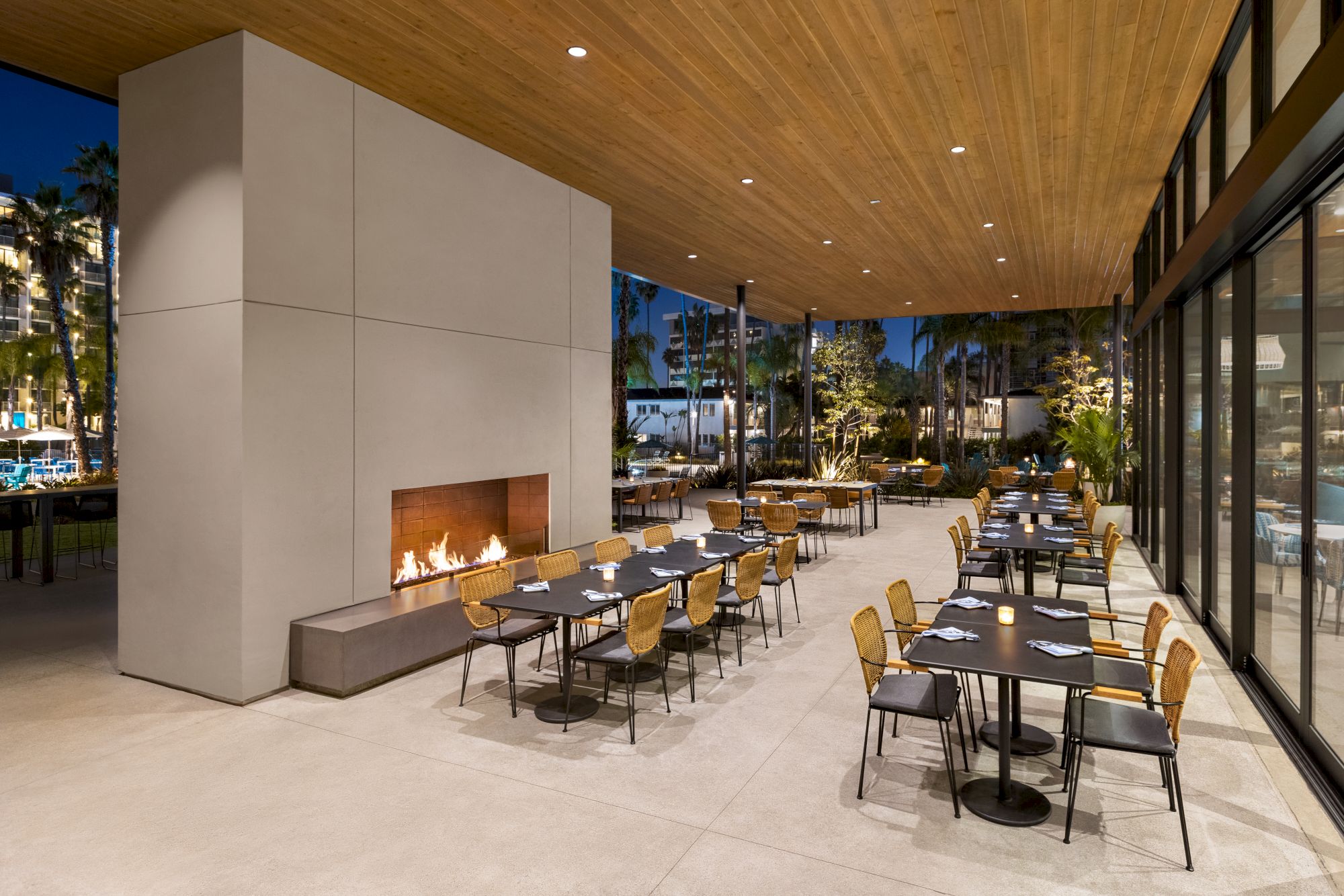 This image shows an outdoor restaurant area with a modern design, featuring a lit fireplace, wooden ceiling, and neatly arranged tables and chairs.