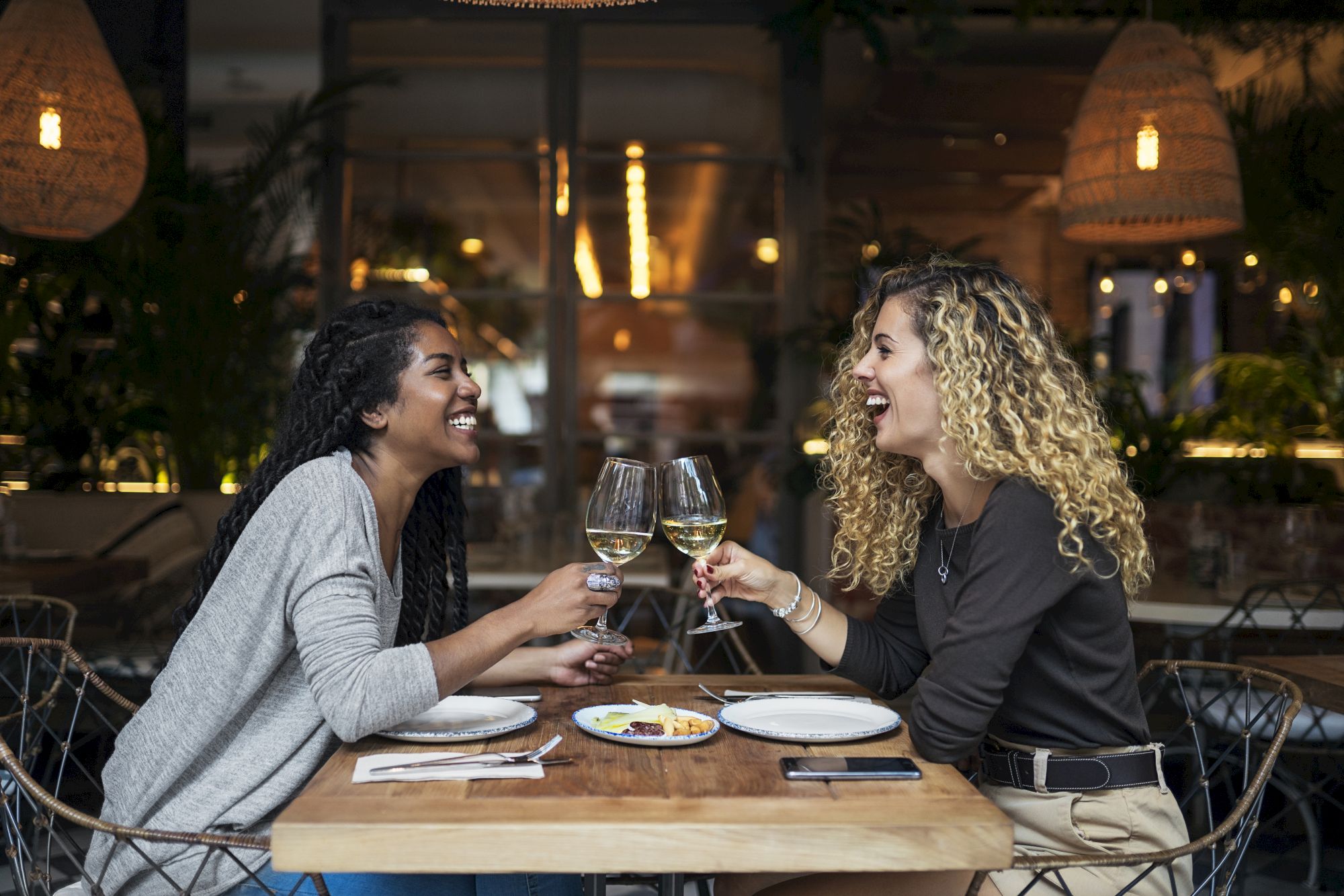 Two people sitting at a restaurant table, smiling and clinking wine glasses, with plates of food in front of them.