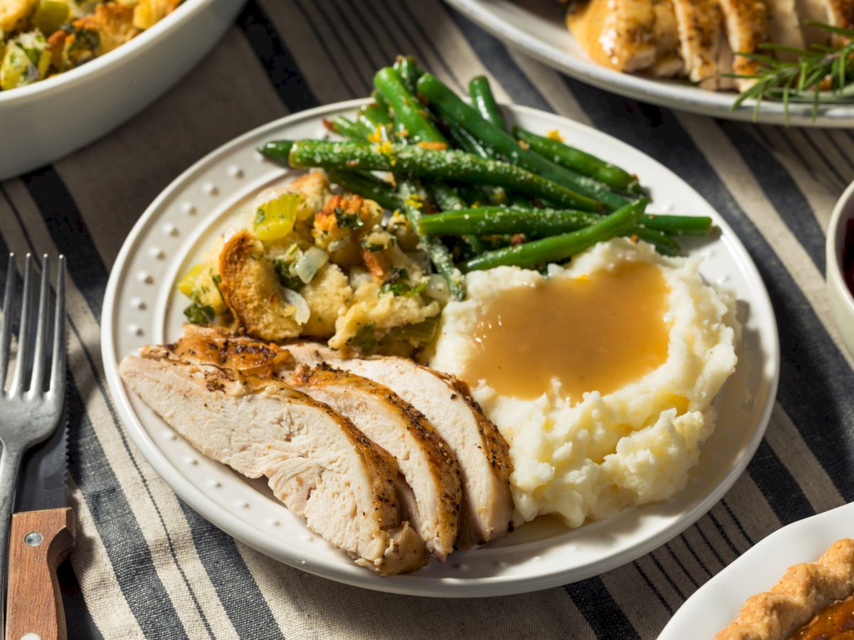 The image shows a plate of sliced turkey, mashed potatoes with gravy, stuffing, and green beans on a striped tablecloth.